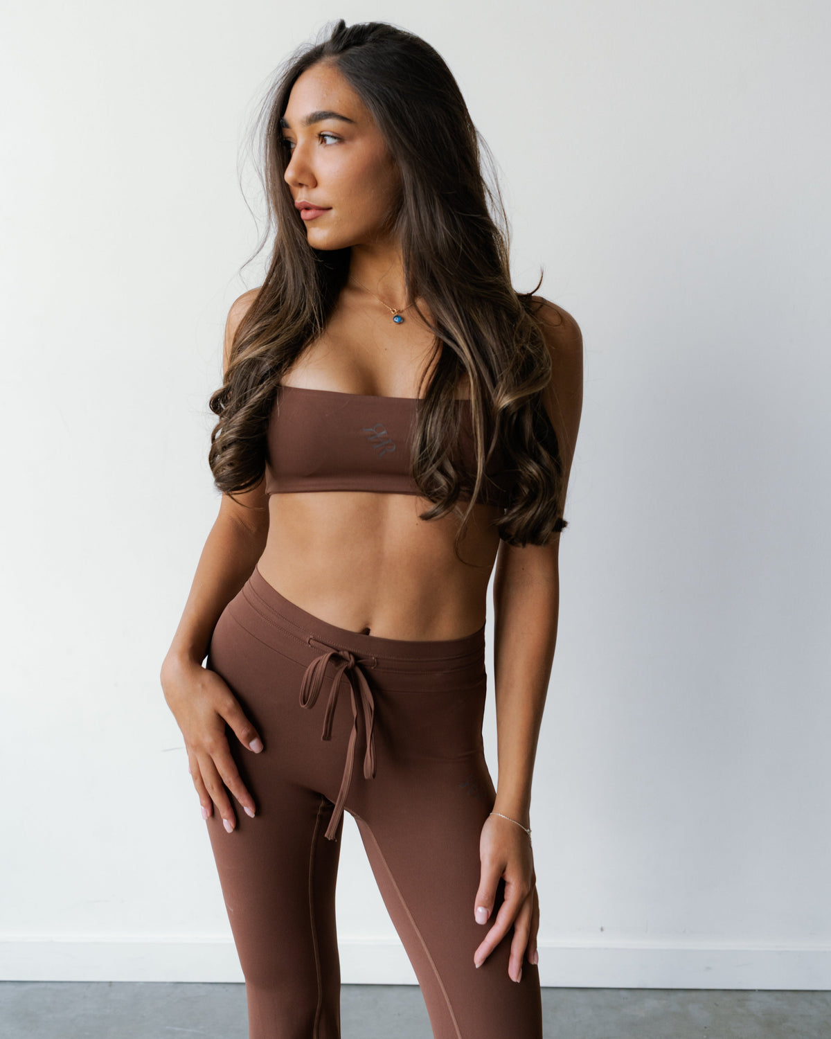 A woman modeling a matching brown activewear set consisting of a strapless bandeau crop top and high-waisted leggings with a drawstring waist, set against a plain white wall.