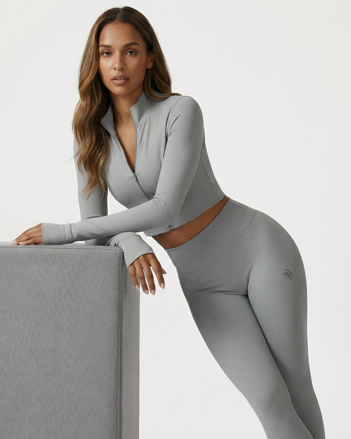 Woman wearing a gray athletic outfit with a visible brand logo, leaning against a gray block on a white background.