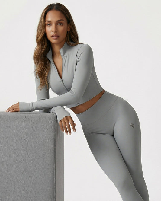 Woman wearing a gray athletic outfit with a visible brand logo, leaning against a gray block on a white background.