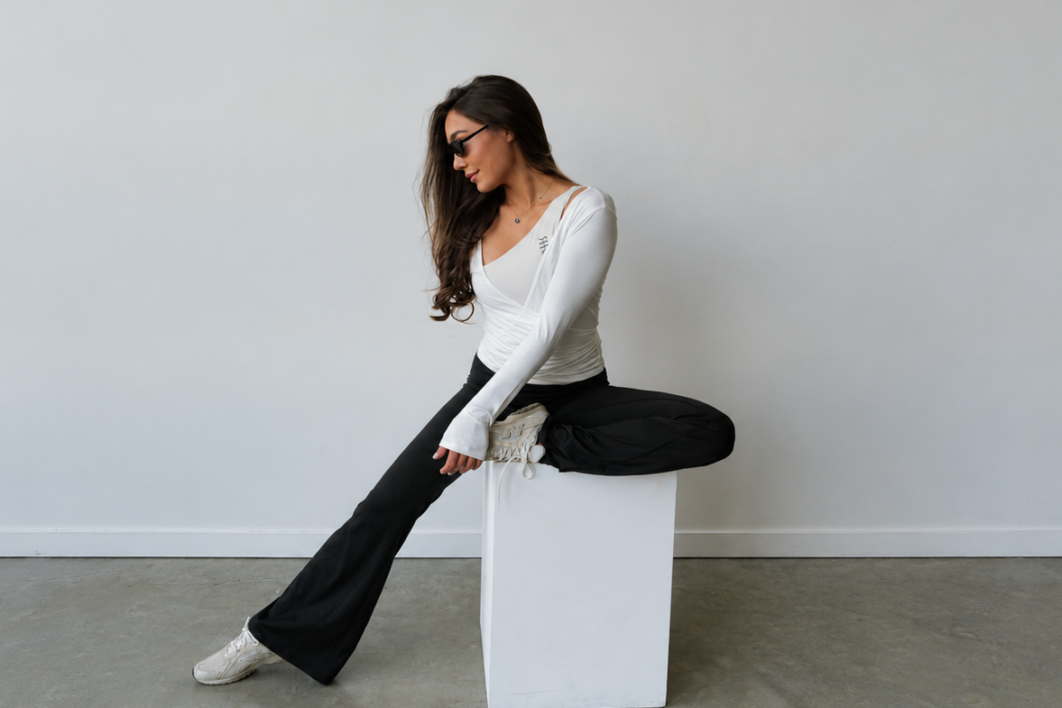 A woman wearing a white long-sleeve top and black flared pants sits cross-legged on a white cube pedestal in a minimalist studio with a gray concrete floor and white wall.