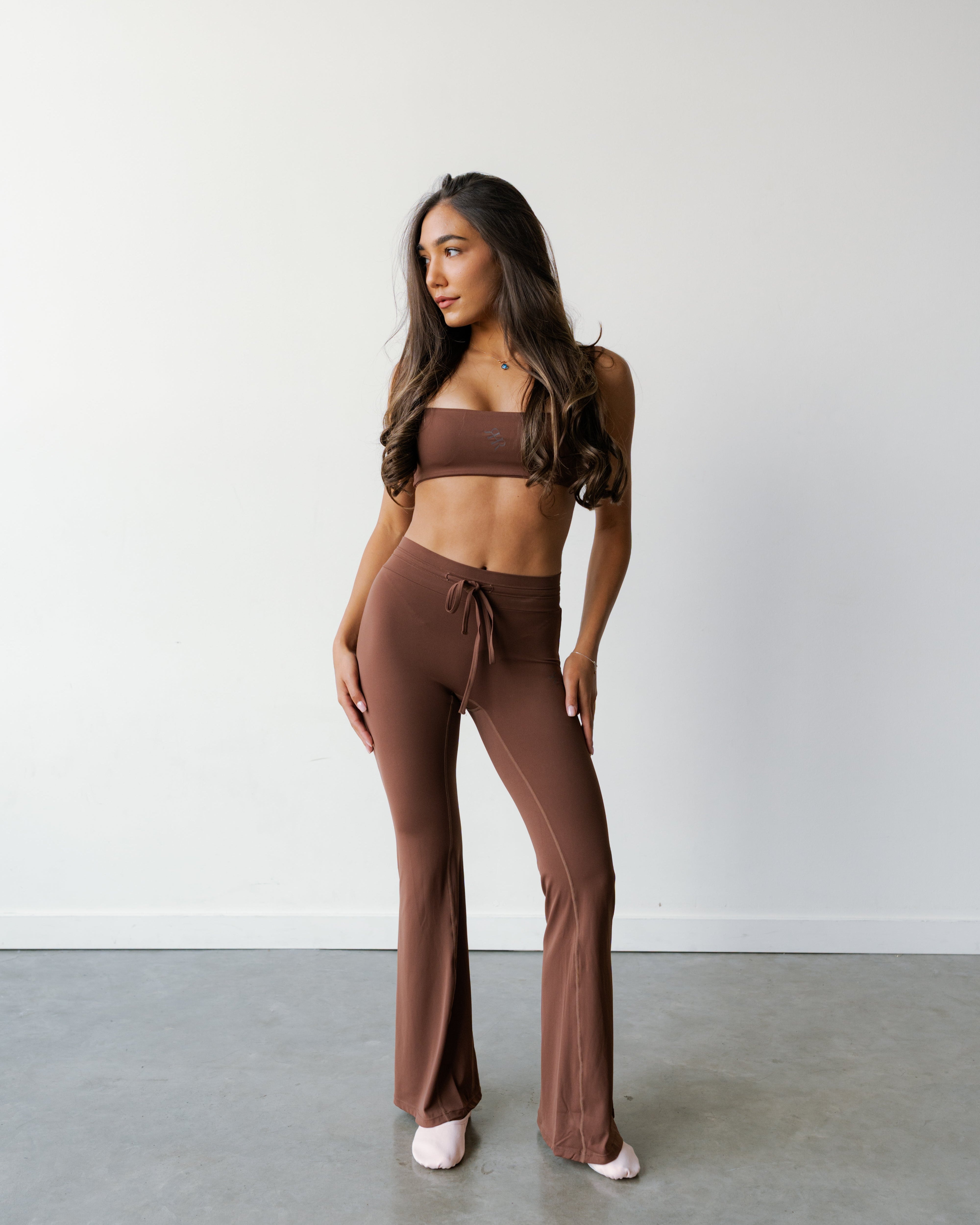 A woman modeling a matching brown lounge set consisting of a bandeau-style crop top and high-waisted drawstring flare pants, standing against a plain white wall and gray concrete floor.