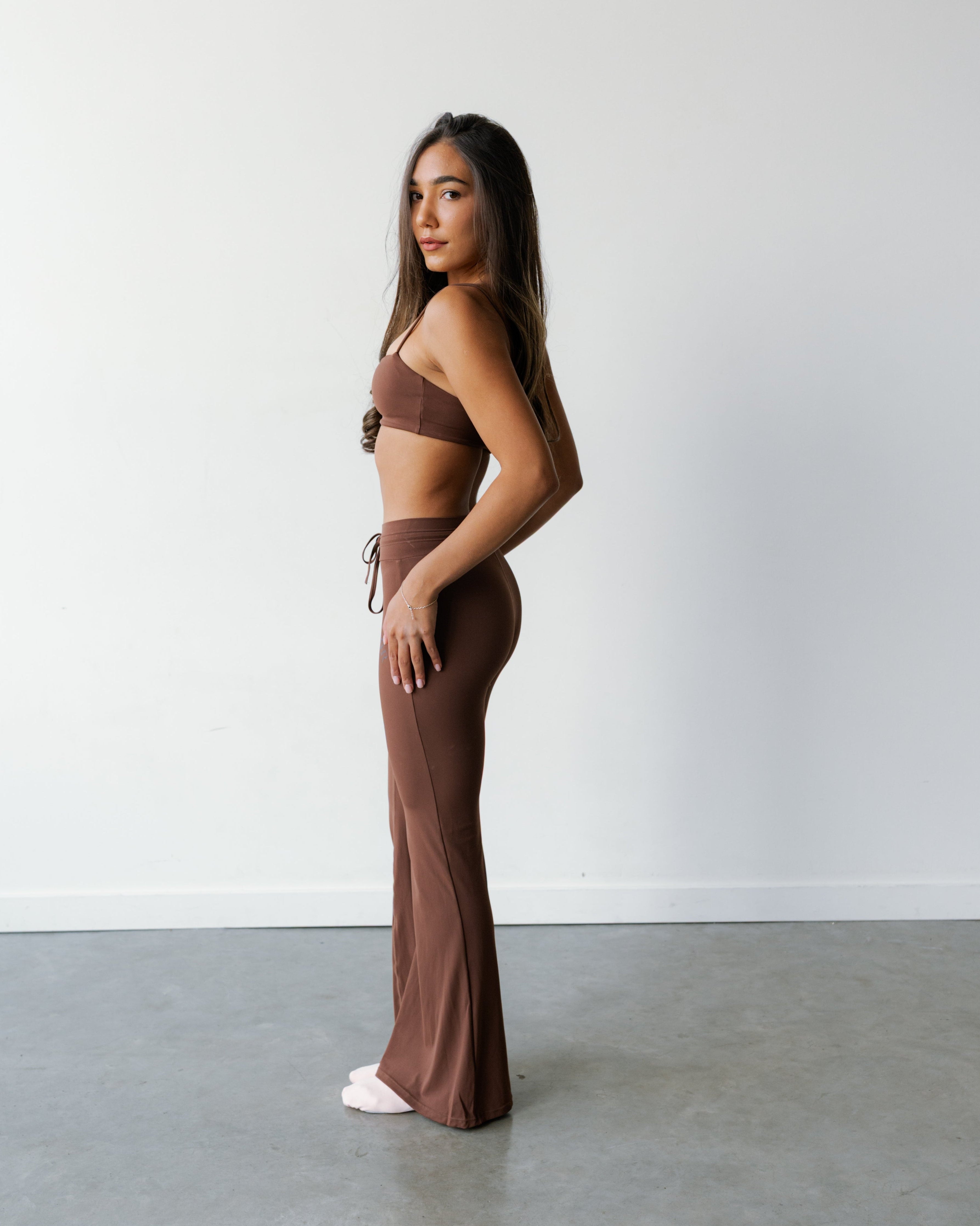 A woman stands in profile wearing a matching brown two-piece outfit consisting of a strapless bandeau top and high-waisted flared pants, photographed against a plain white wall and gray concrete floor.