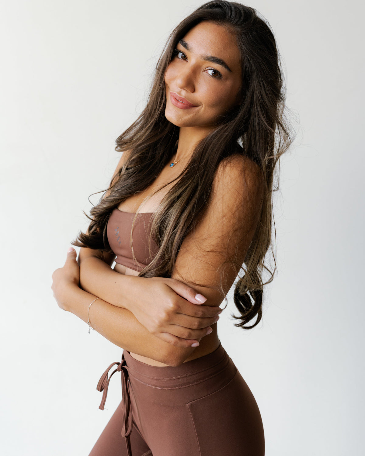 A woman posing in a matching brown activewear set consisting of a sports bra and high-waisted leggings, set against a plain light background.