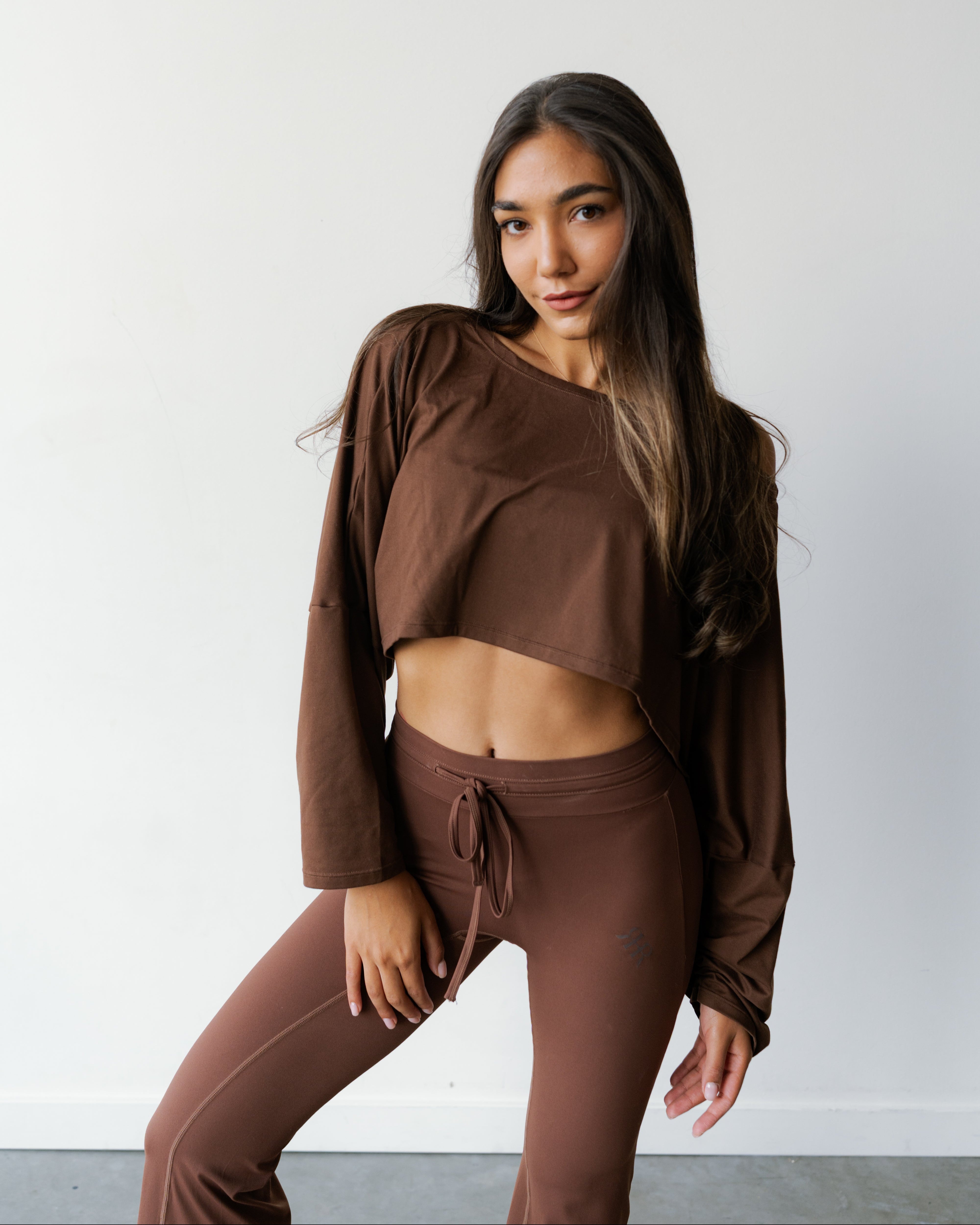 A woman modeling a matching brown activewear set consisting of a long-sleeve cropped top and high-waisted drawstring leggings, set against a plain light gray wall.