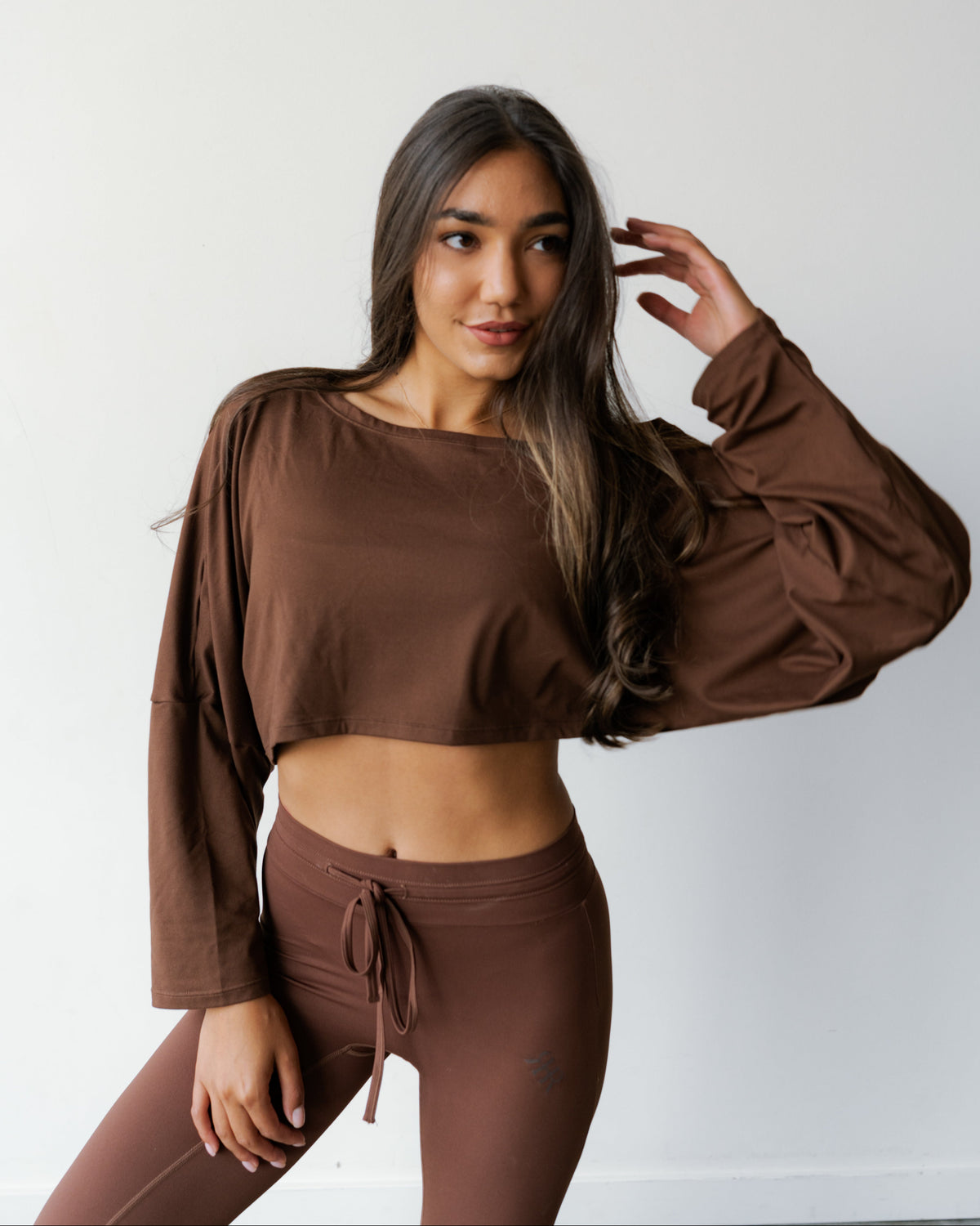 A woman modeling a matching brown lounge set consisting of a cropped long-sleeve top and high-waisted drawstring leggings, posed against a plain white wall.
