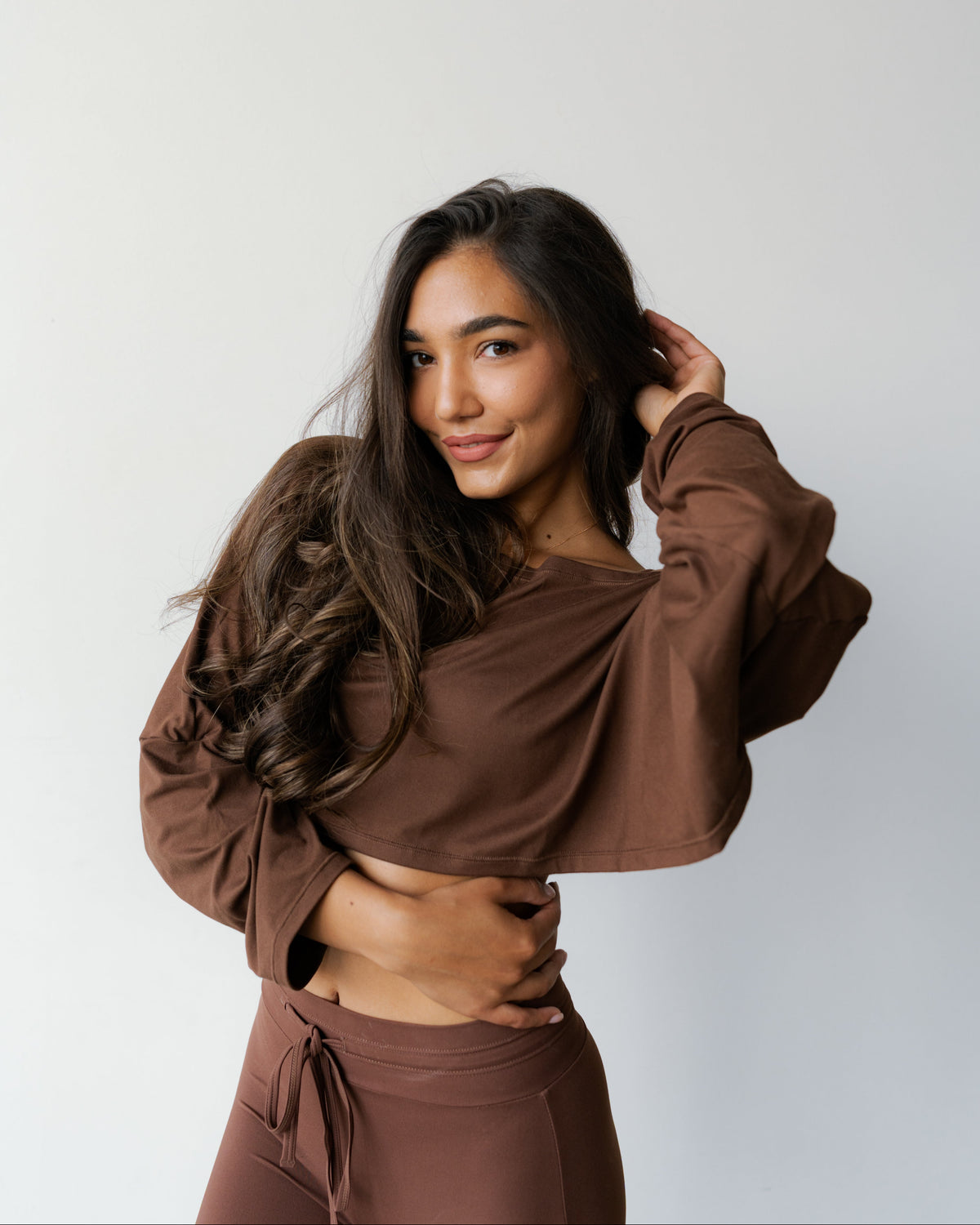 A woman modeling a matching brown two-piece loungewear set, featuring a long-sleeve crop top and high-waisted drawstring pants, against a plain light gray background.