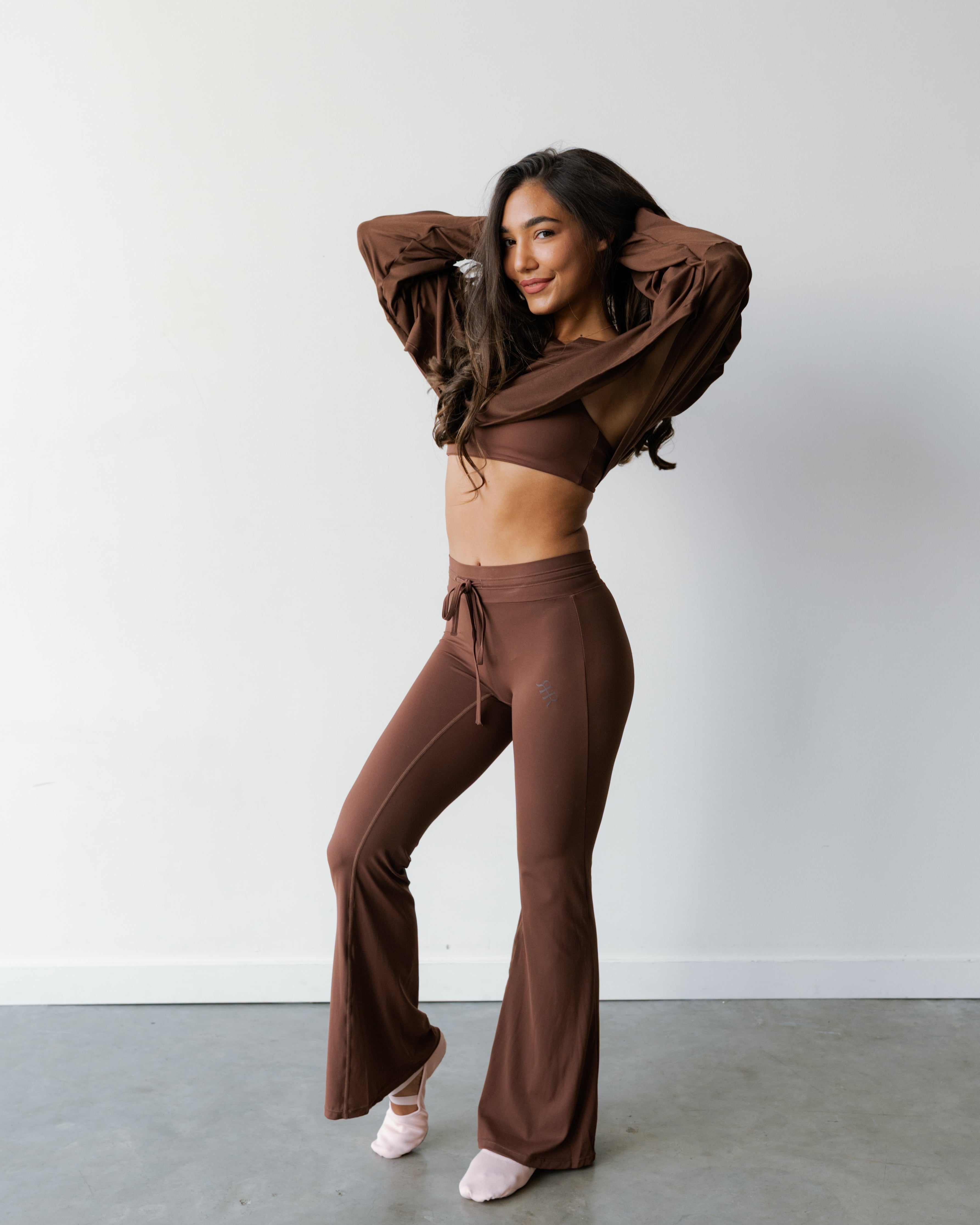 A woman modeling a matching brown lounge set consisting of a long-sleeve crop top and high-waisted flared pants, standing in a minimalist studio with a white wall and gray concrete floor.
