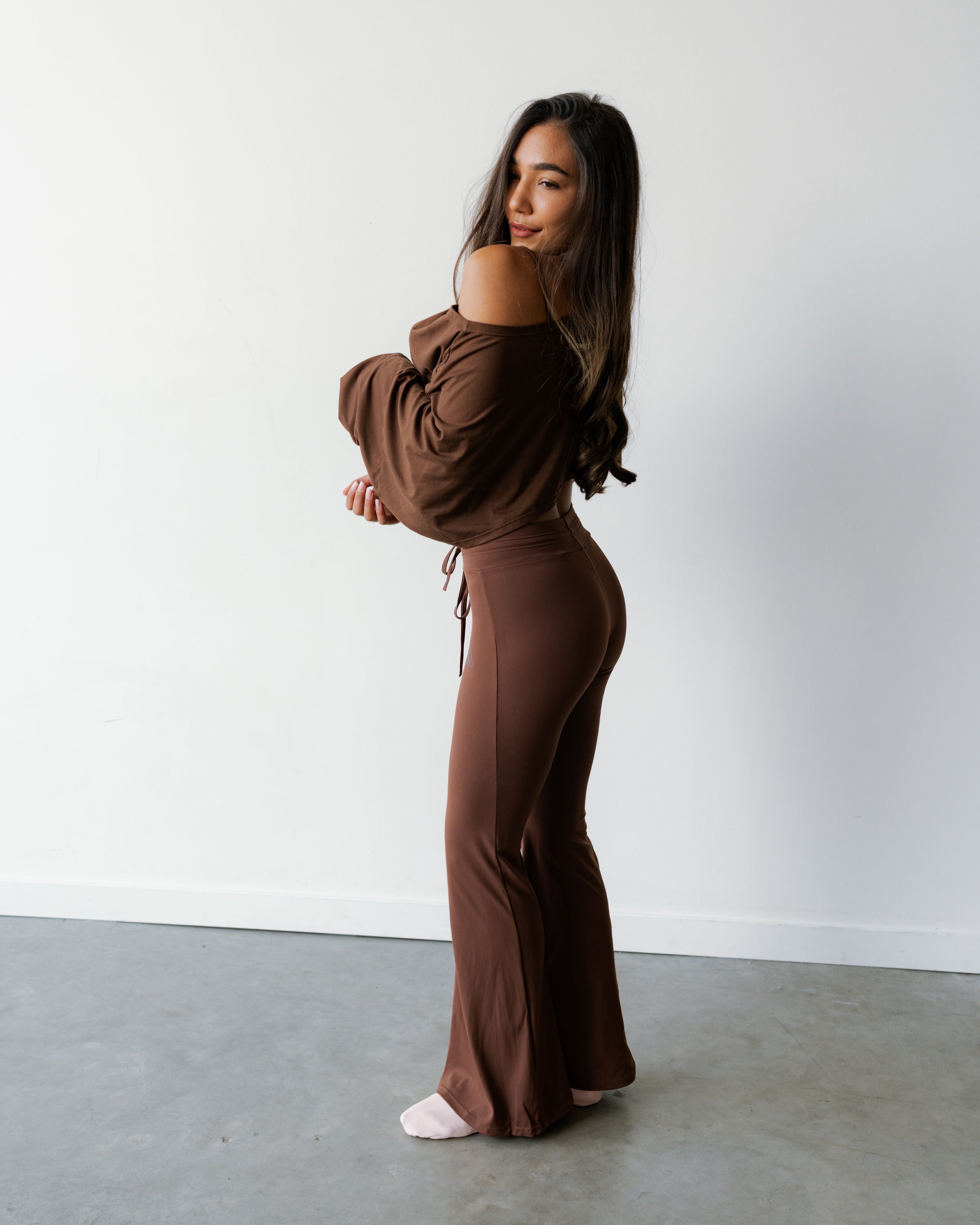 A woman modeling a matching brown off-the-shoulder long-sleeve top and high-waisted flared pants set, standing in a minimalist studio with a white wall and gray concrete floor.