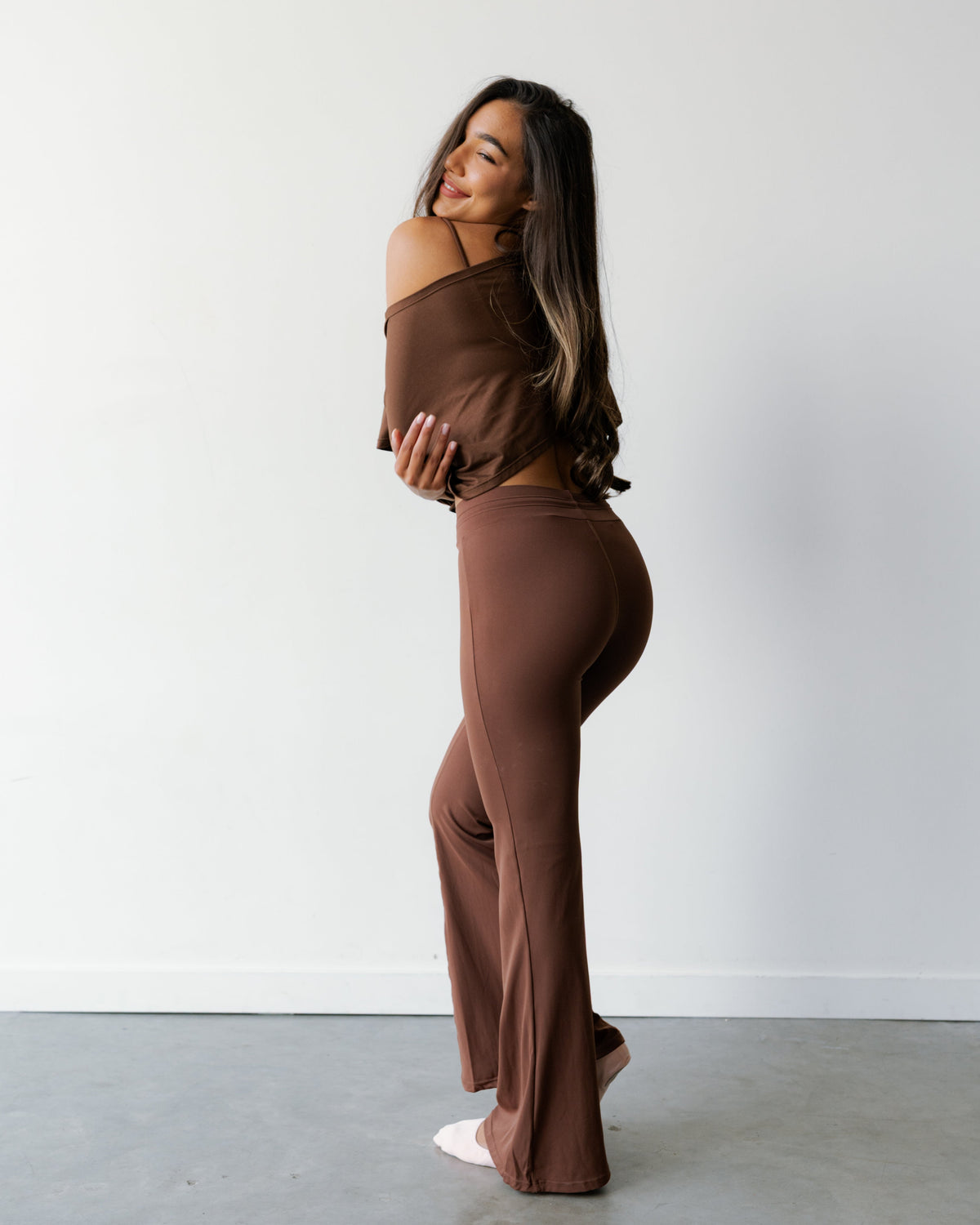 A woman modeling a matching two-piece lounge set in a solid brown color, featuring an off-the-shoulder long-sleeve top and high-waisted flared pants, standing against a minimalist white wall and gray concrete floor.
