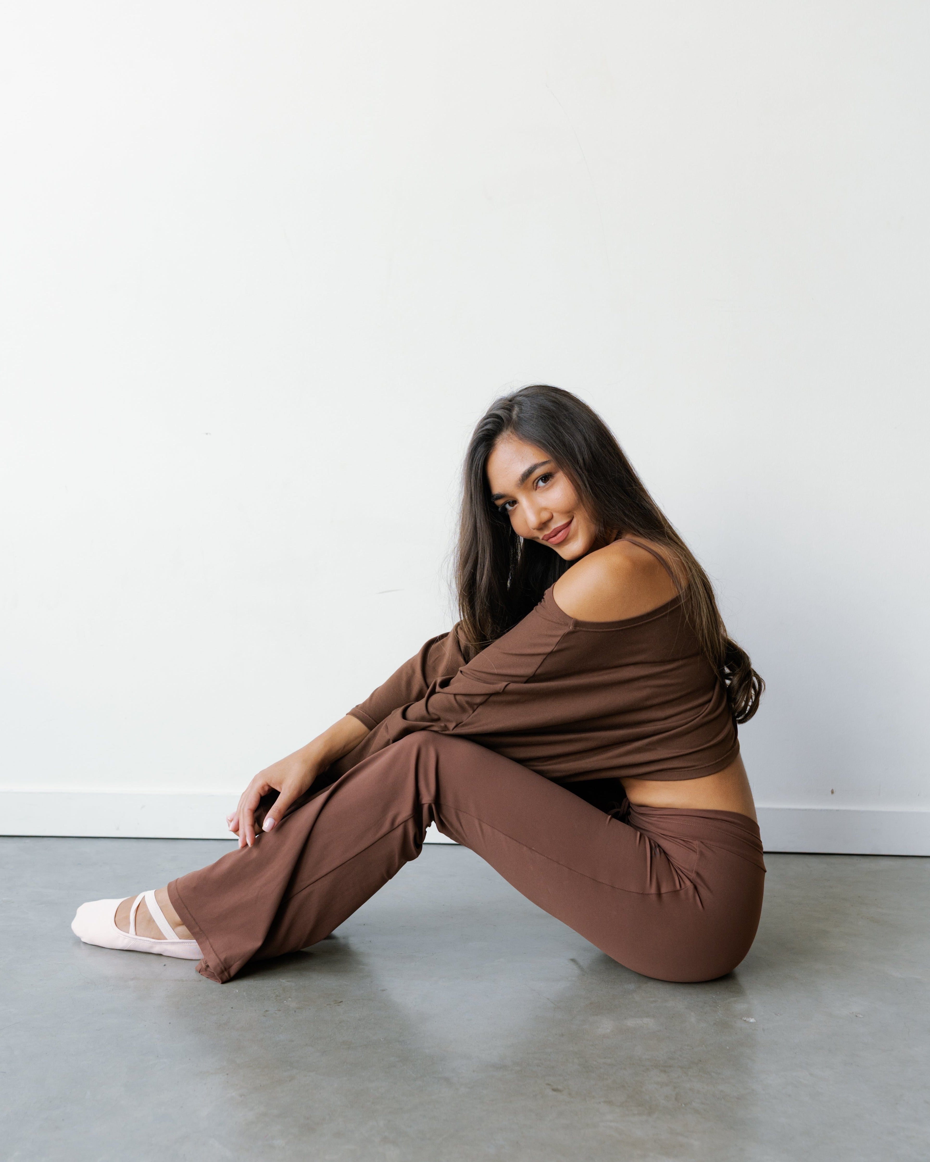 A woman sits on a concrete floor against a white wall, wearing a matching brown off-the-shoulder long-sleeve top and wide-leg pants, with white ballet slippers.
