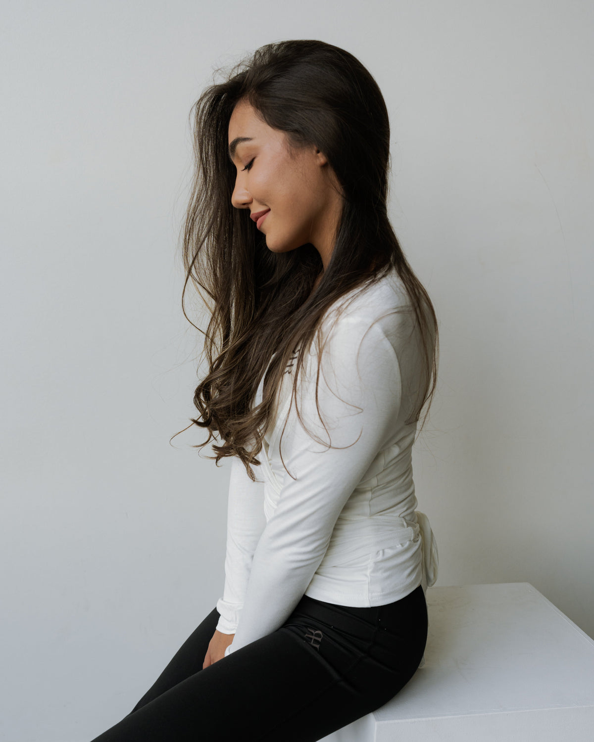 A woman with long dark hair sits on a white block, wearing a fitted white long-sleeve top and black pants, photographed against a plain light gray wall.