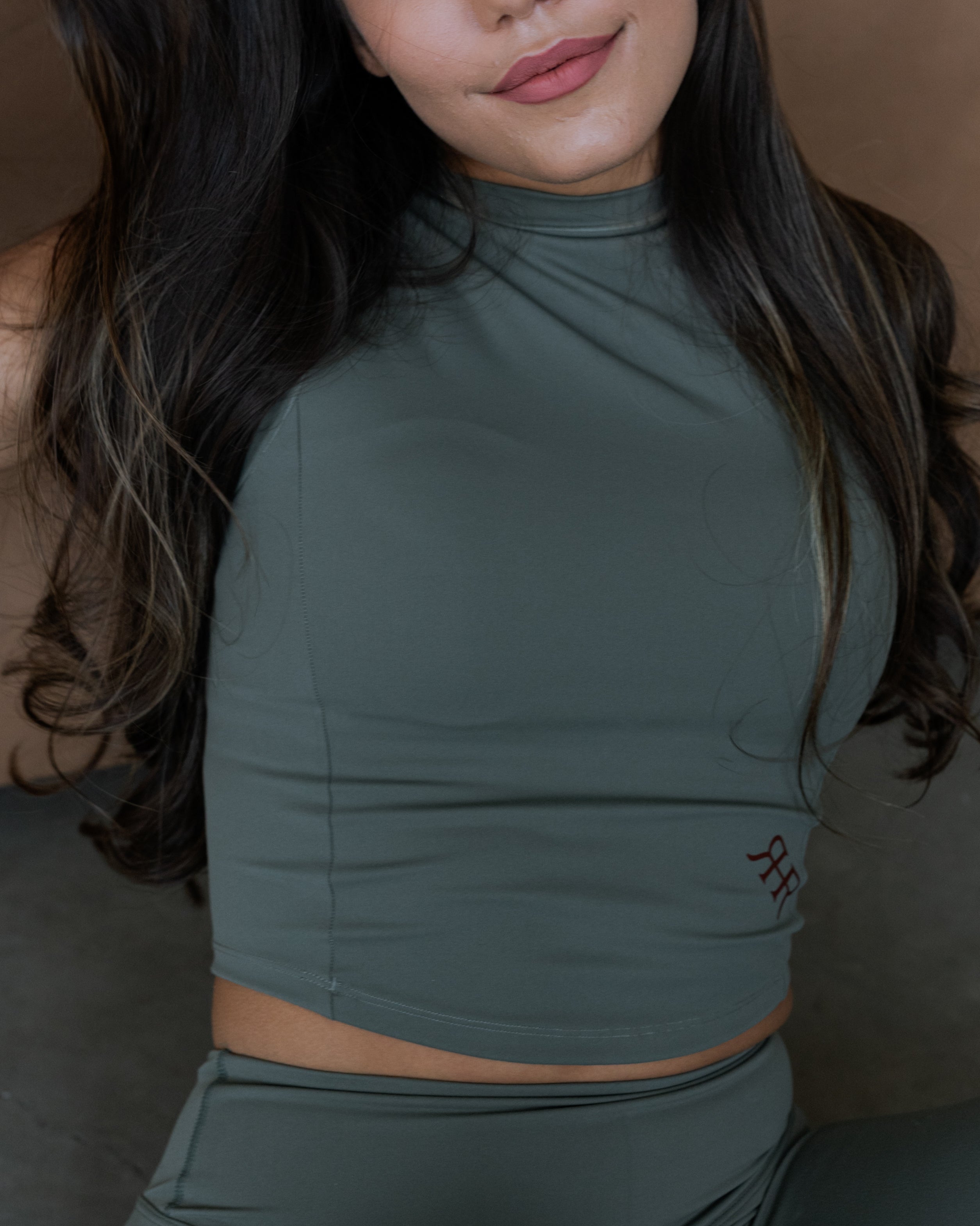A close-up of a woman wearing a dark sage green athletic crop top and matching leggings, featuring a small embroidered logo on the lower right side of the top.