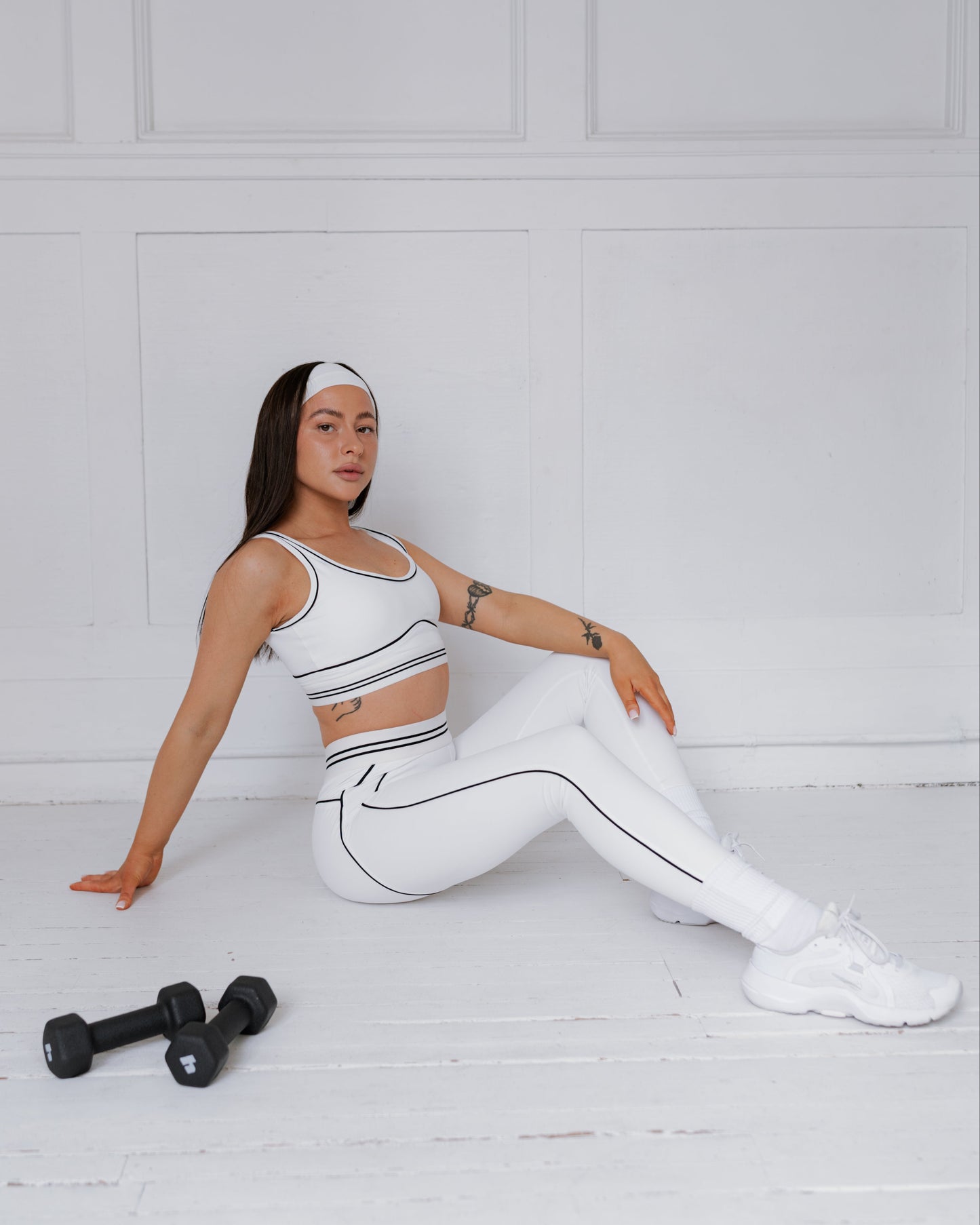 Woman in white athletic wear sitting on a white floor with black dumbbells.
