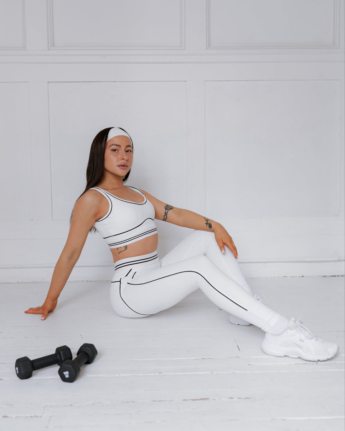 Woman in white athletic wear sitting on a white floor with black dumbbells.
