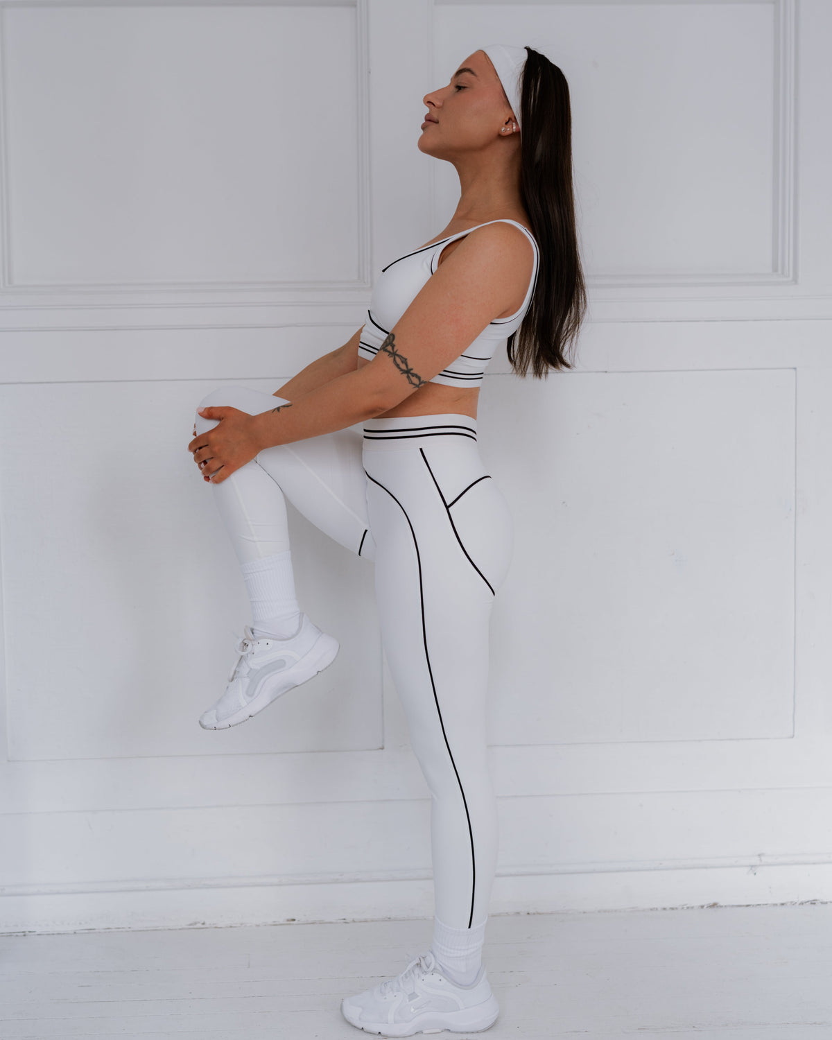 Woman in a white and black athletic outfit stretching against a white wall.