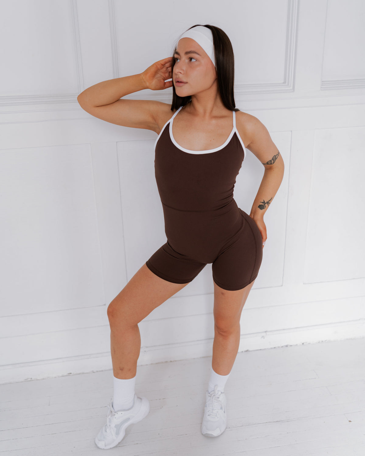 Woman wearing a brown bodysuit with white accents against a white wall.
