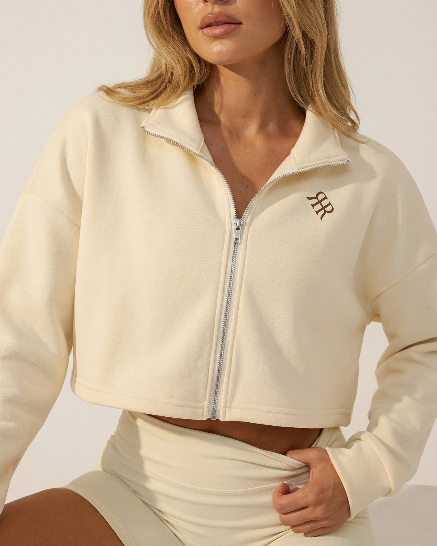 Woman wearing a cream-colored athletic jacket with a logo on a neutral background