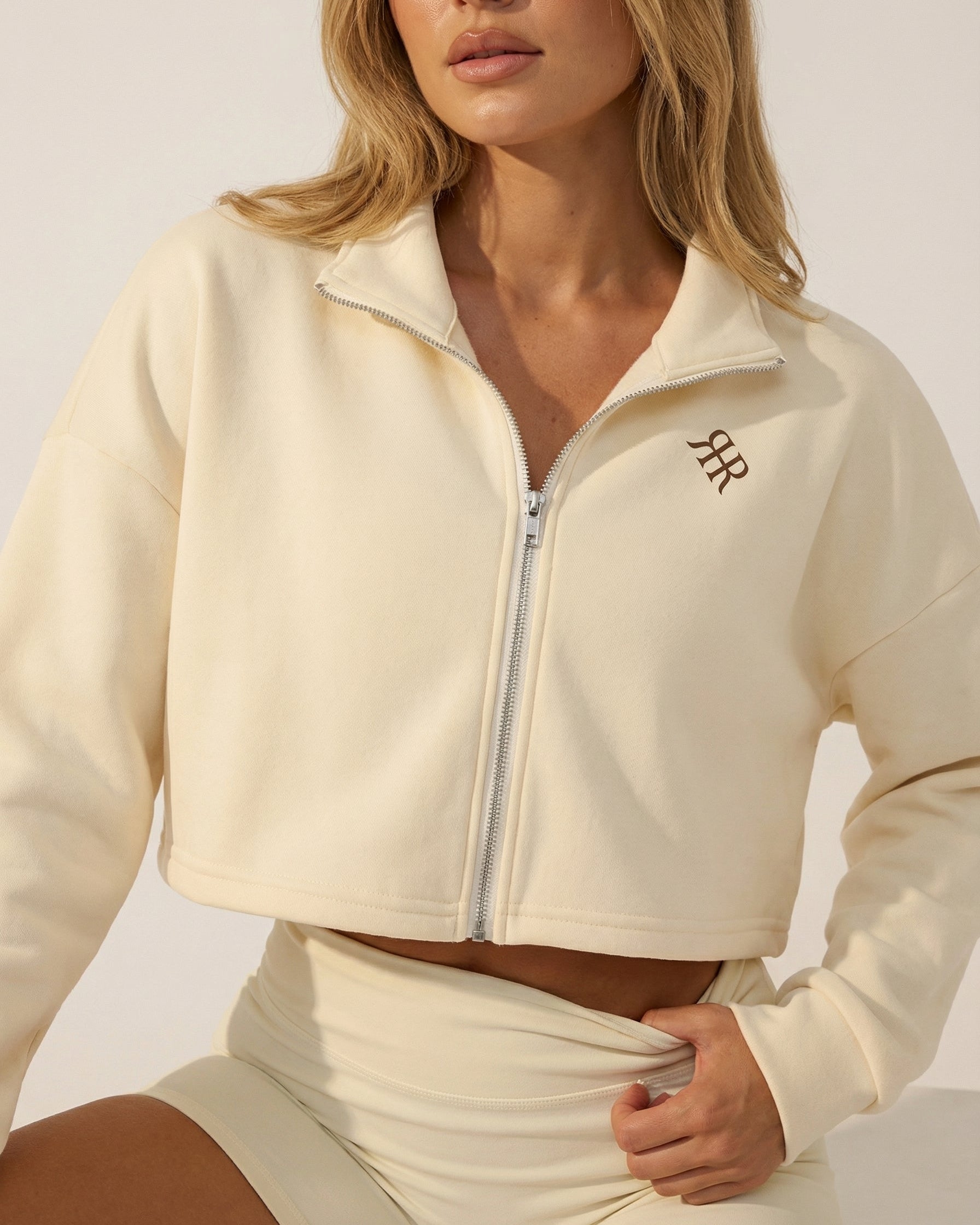 Woman wearing a cream-colored athletic jacket with a logo on a neutral background