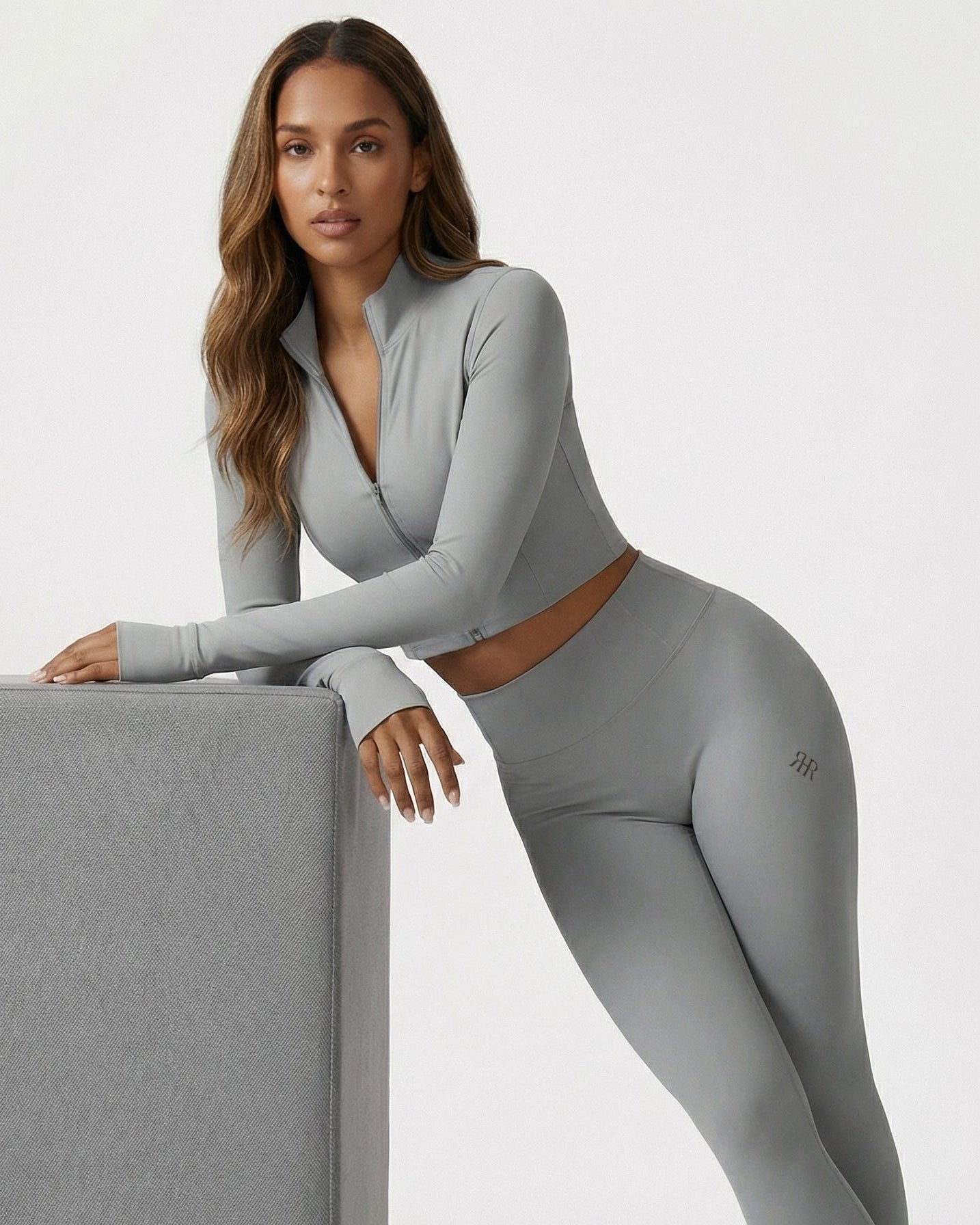 Woman wearing a gray athletic outfit with a visible brand logo, leaning against a gray block on a white background.